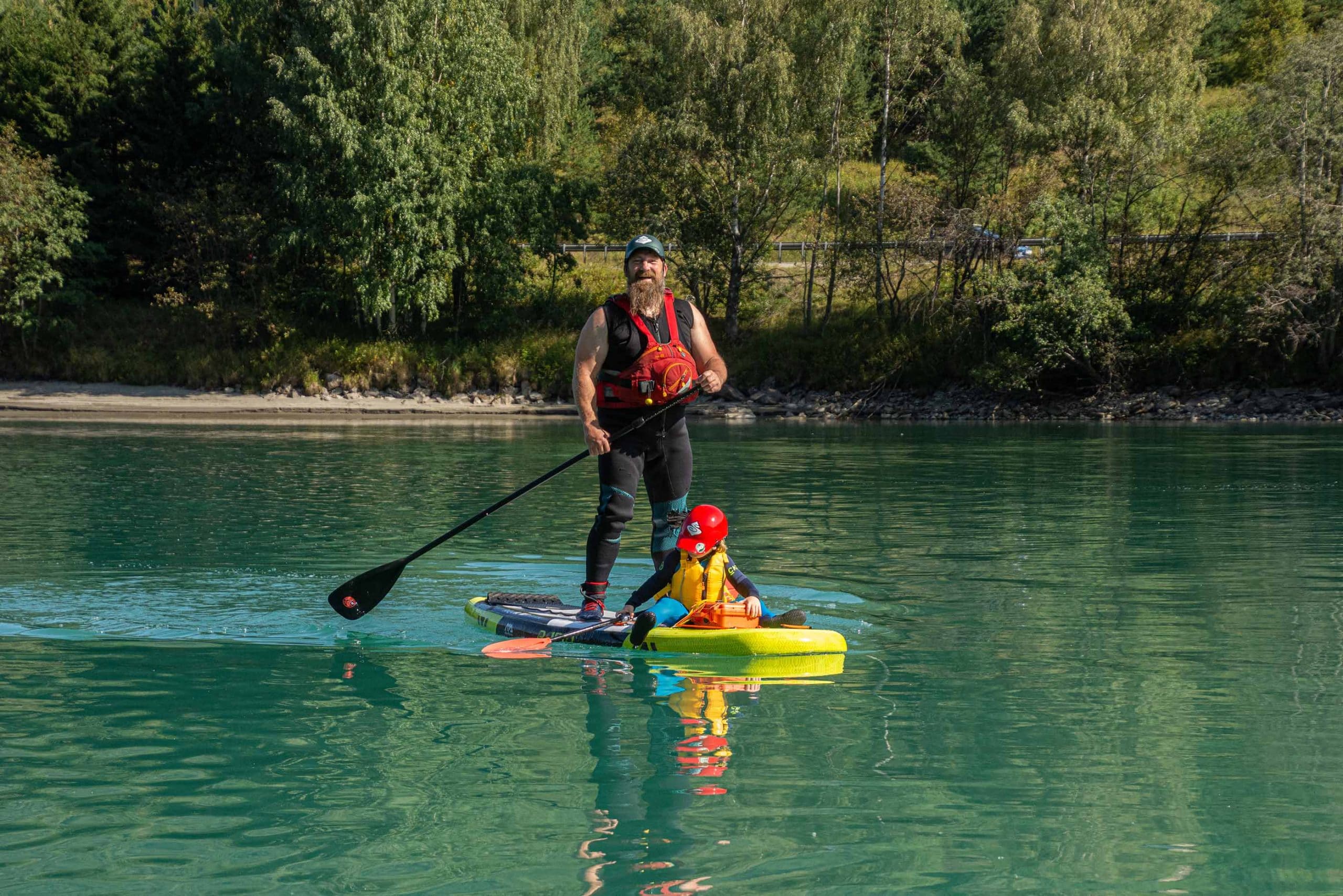 En mann stående på SUP brett med et lite barn sittende foran på brettet